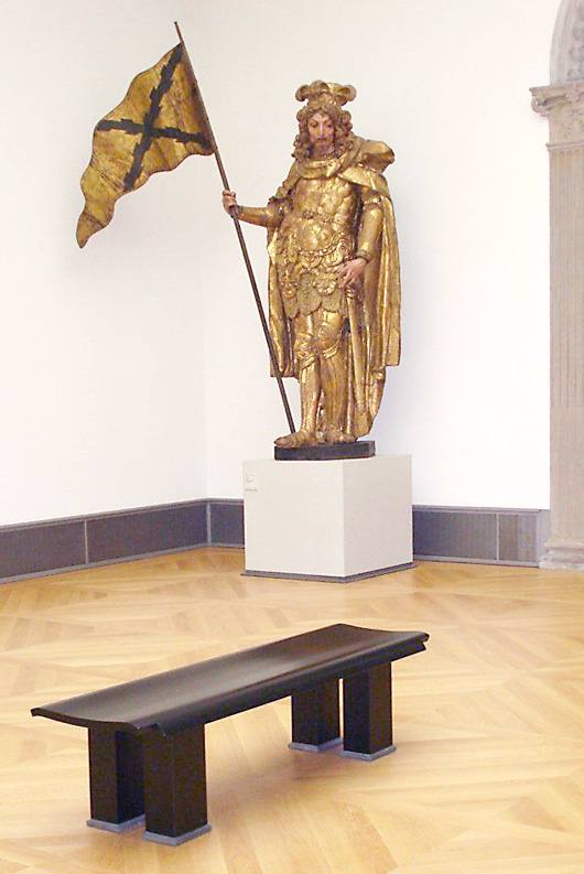 Dunkle Sitzbank mit geschwungener Sitzfläche vor einer vergoldeten Fahnenträger-Skulptur im Bode-Museum