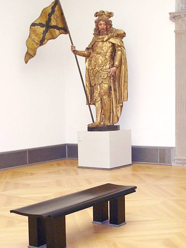 Dunkle Sitzbank mit geschwungener Sitzfläche vor einer vergoldeten Fahnenträger-Skulptur im Bode-Museum