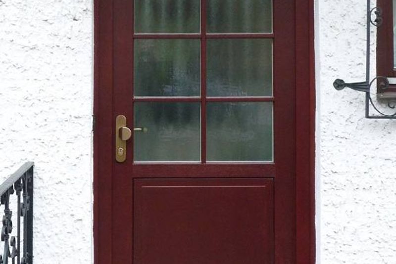 Rot lackierte Holzhaustür mit Sprossenverglasung und Sicherheitsbeschlag in Messing an einem weiß verputzten Haus