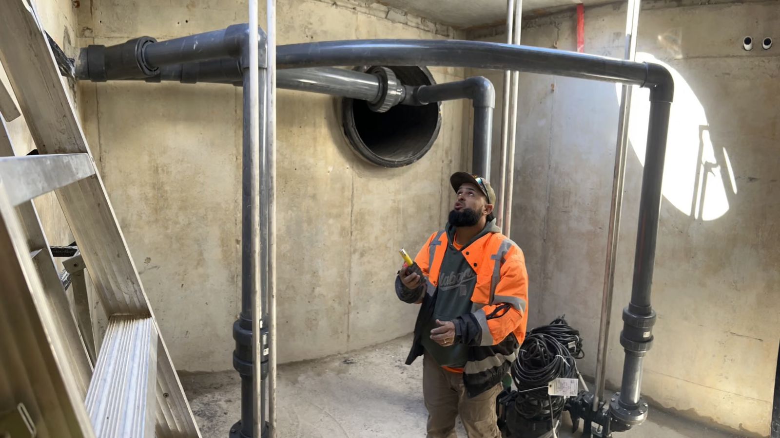 A worker in an orange safety jacket looks up at gray SubAir piping and valves inside a concrete equipment vault