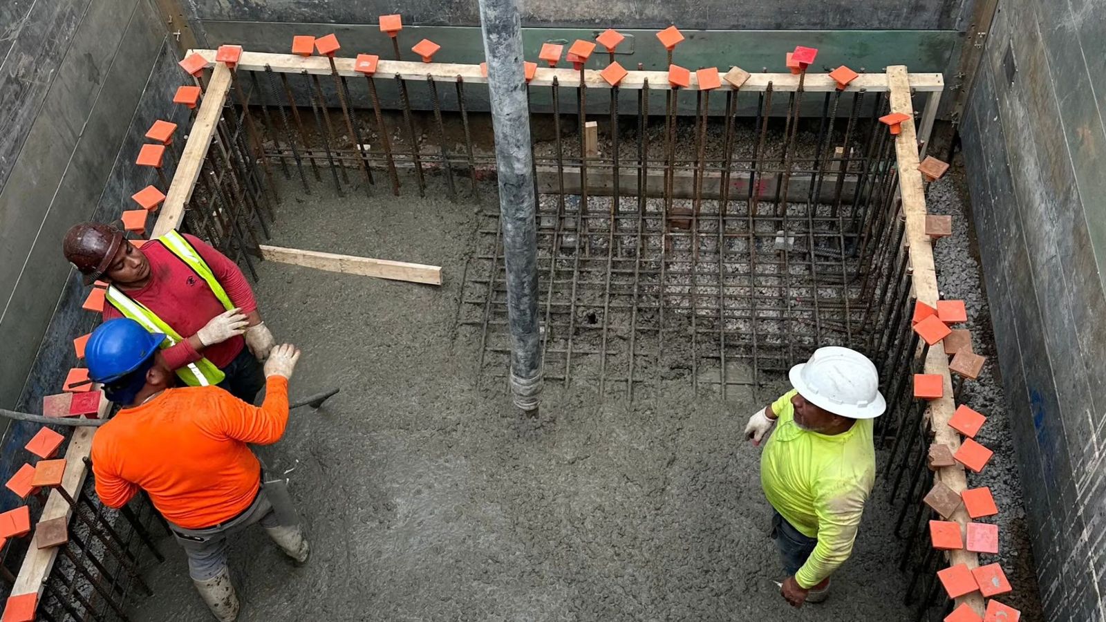 Three workers place concrete inside a rebar cage at the bottom of a shored excavation pit