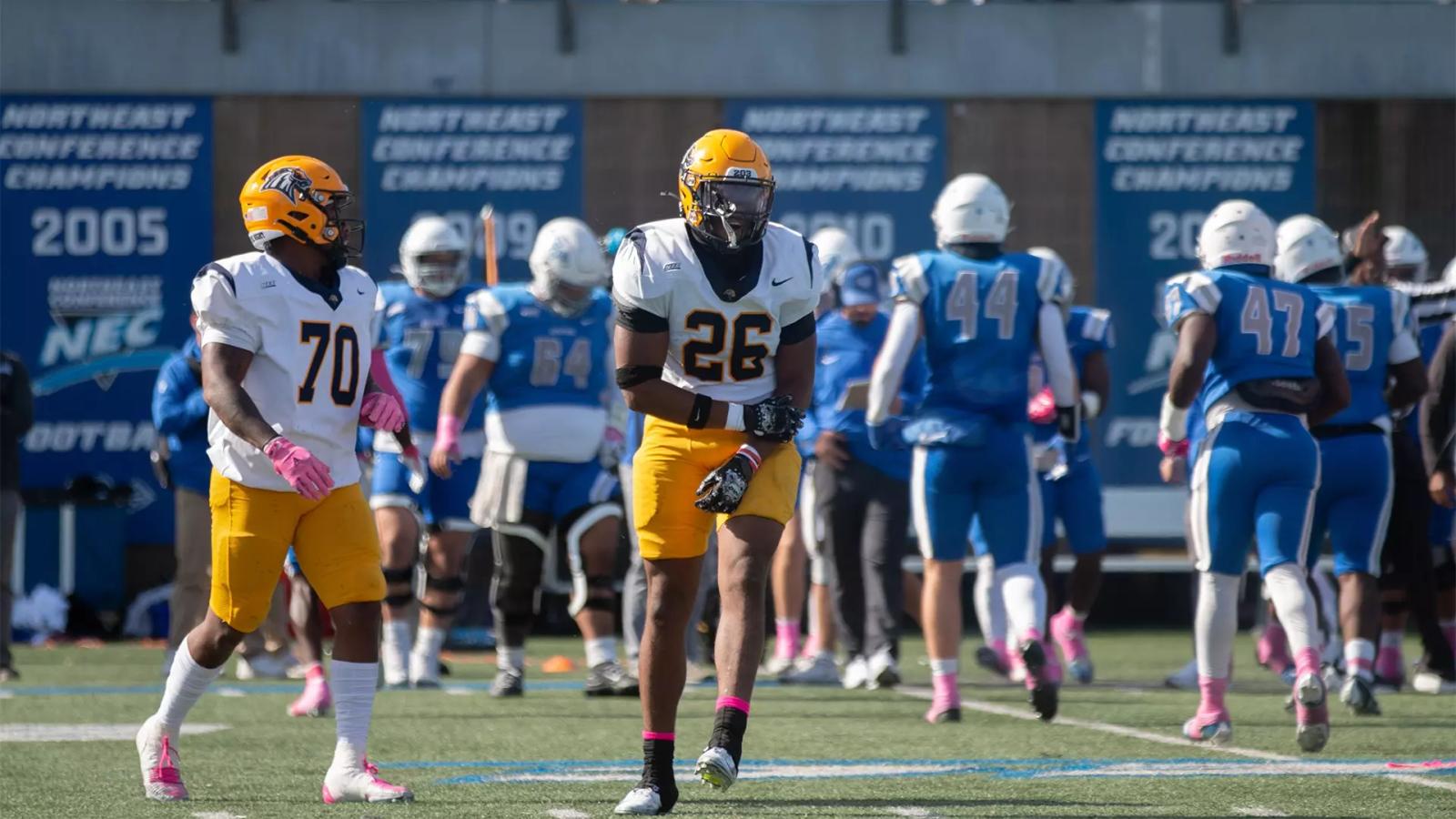New Haven players No. 70 and No. 26, in white jerseys and gold pants, on the field at Central Connecticut's Arute Field with Blue Devils players in blue behind them on Oct. 24, 2026