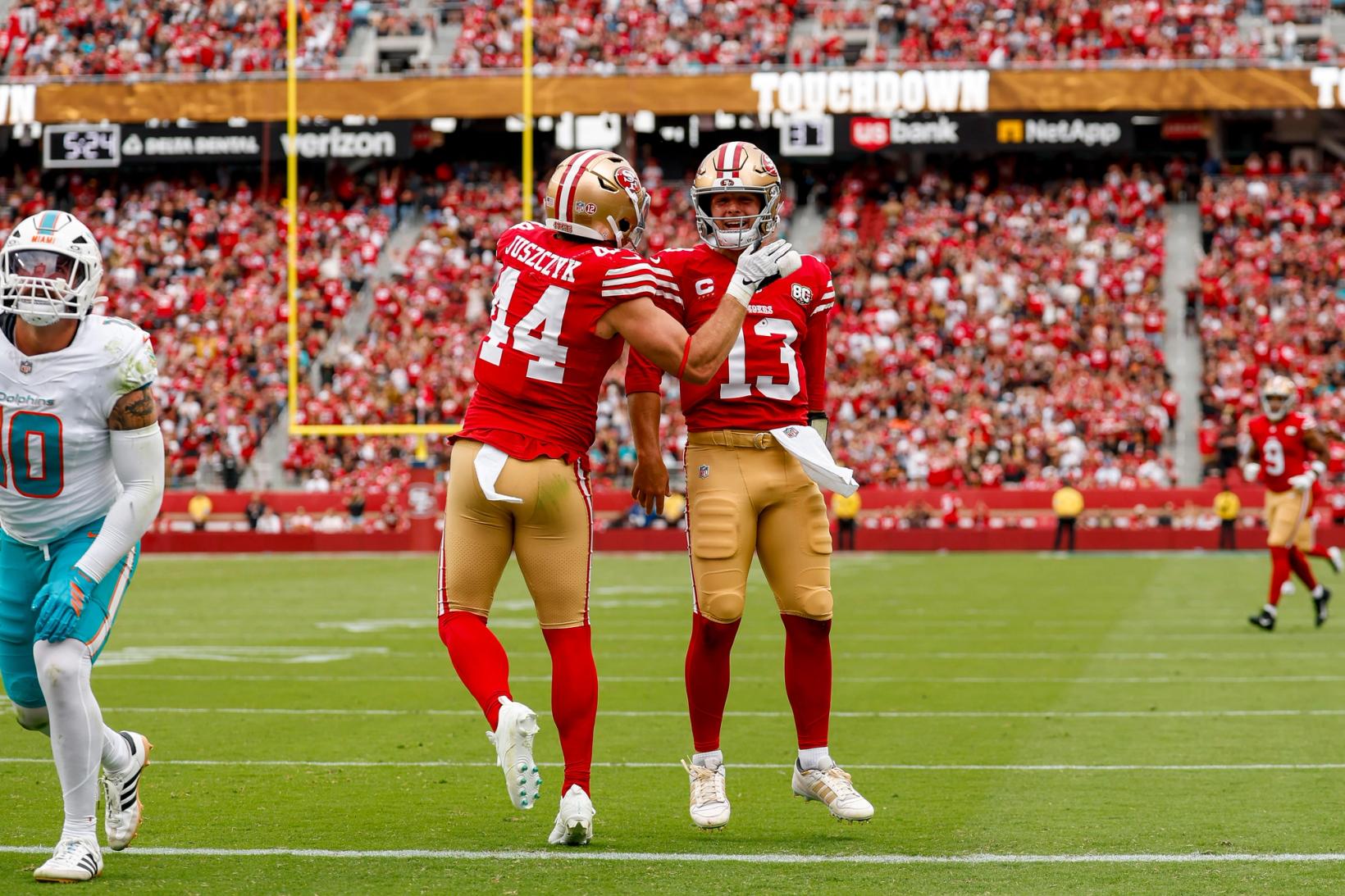 San Francisco 49ers fullback Kyle Juszczyk (44) and quarterback Brock Purdy (13) celebrate on the field at Levi's Stadium as a Miami Dolphins player walks away and the ribbon board behind them reads Touchdown
