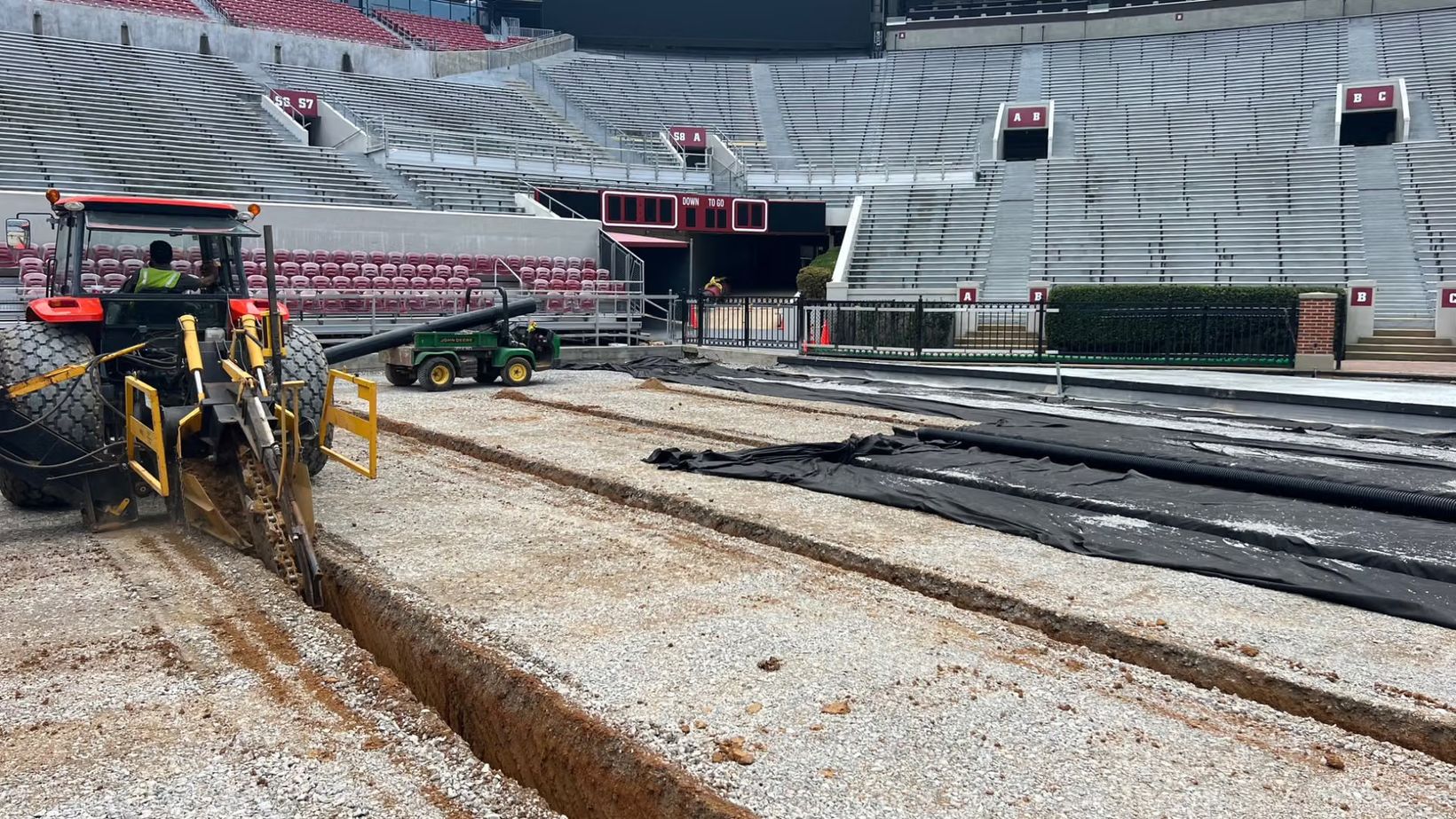 A trenching machine cuts drainage lines through the gravel base of the Bryant-Denny Stadium field during the 2023 rebuild, with rolls of black drain pipe and fabric laid out beside it