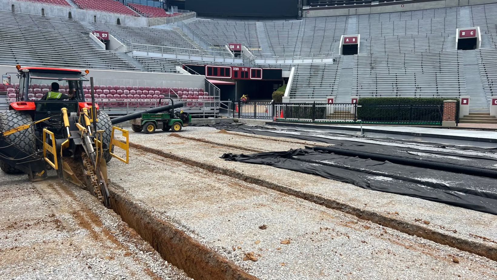 A trenching machine cuts drainage lines through the gravel base of the Bryant-Denny Stadium field during the 2023 rebuild, with rolls of black drain pipe and fabric laid out beside it
