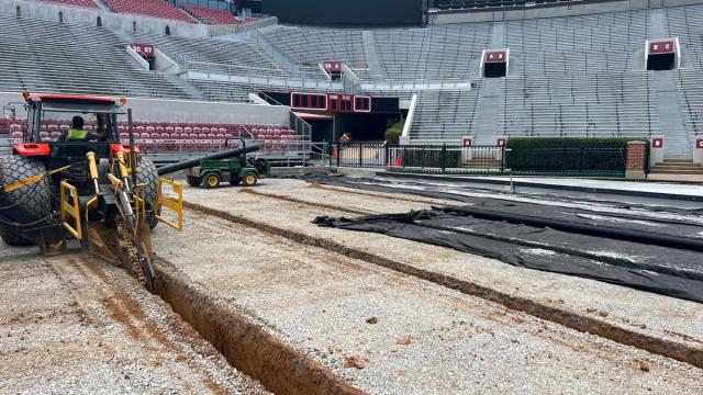 A trenching machine cuts drainage lines through the gravel base of the Bryant-Denny Stadium field during the 2023 rebuild, with rolls of black drain pipe and fabric laid out beside it