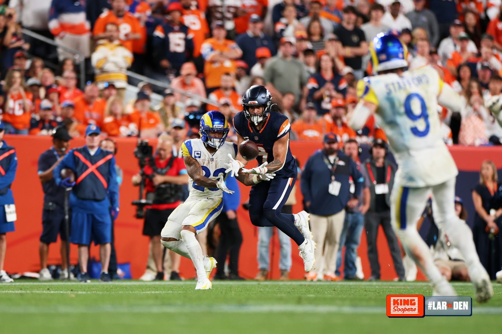 A Denver Broncos defender catches an interception in front of a Los Angeles Rams player as Rams quarterback Matthew Stafford (9) trails the play in the foreground at Empower Field at Mile High.