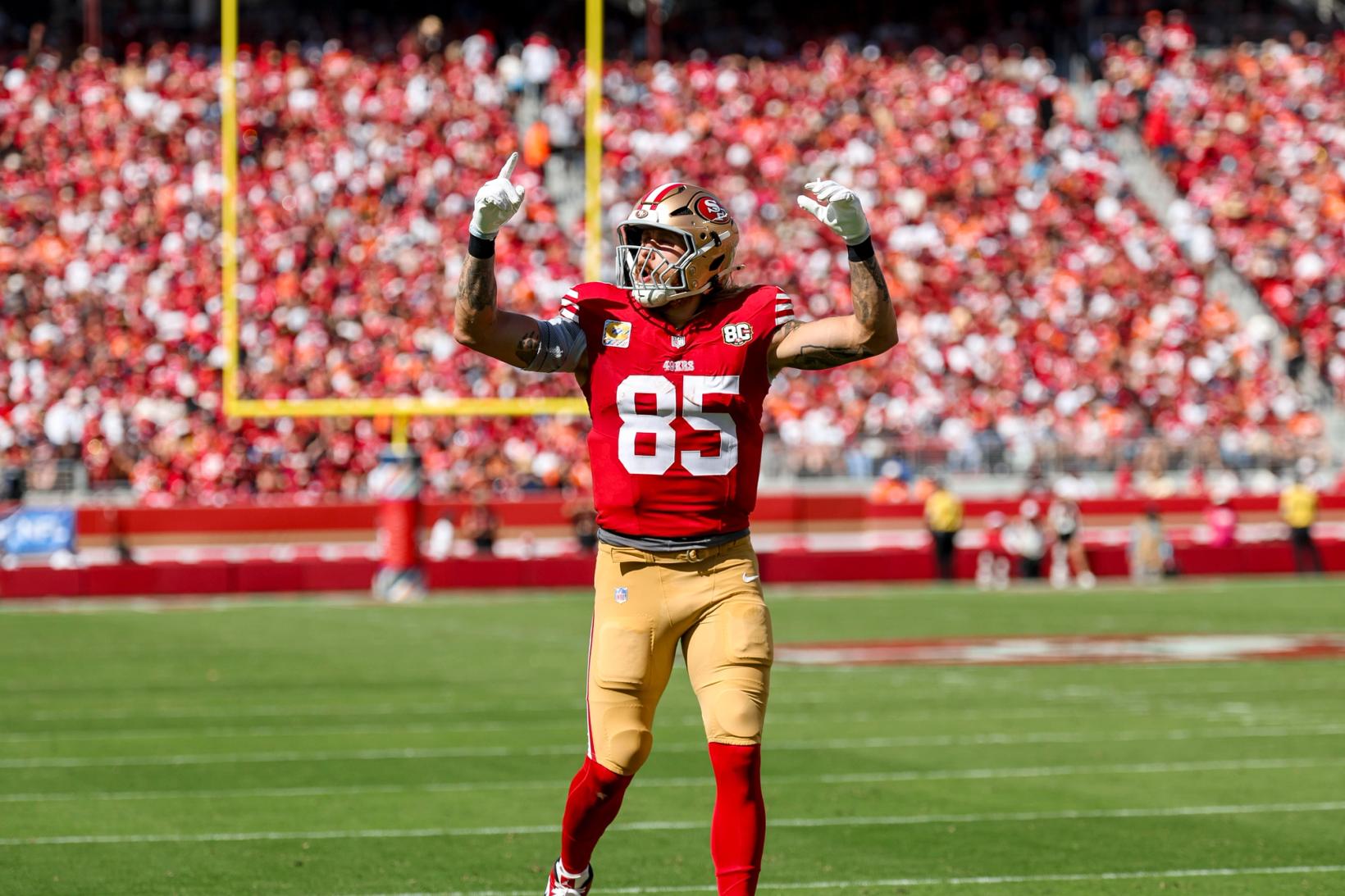 San Francisco 49ers tight end George Kittle (85) raises both arms toward a packed crowd in red at Levi's Stadium, with a goalpost behind him