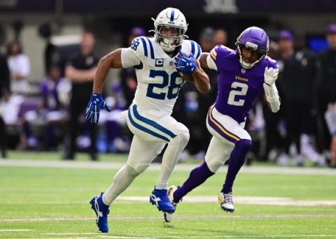 Indianapolis Colts running back Jonathan Taylor, wearing No. 28 and a captain's patch, carries the ball upfield as a Minnesota Vikings defender wearing No. 2 chases him.