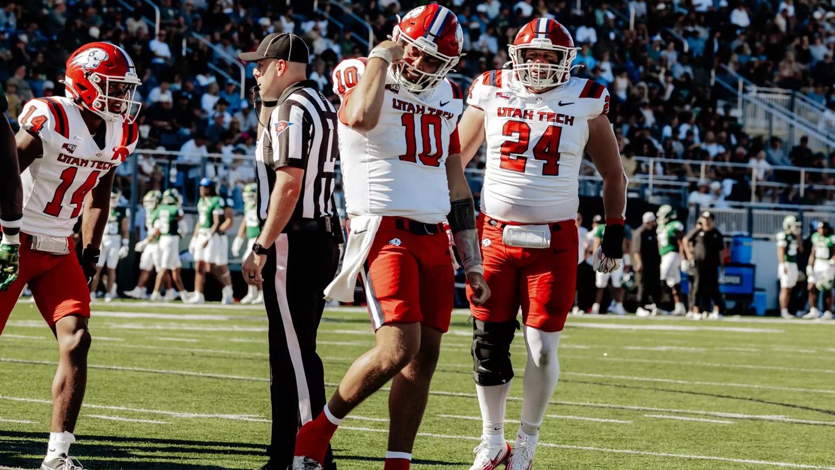 Utah Tech players in white jerseys and red helmets, including Nos. 10, 24 and 14, walk the field beside an official as Cal Poly players in green stand on the sideline at Spanos Stadium, Oct. 24, 2026