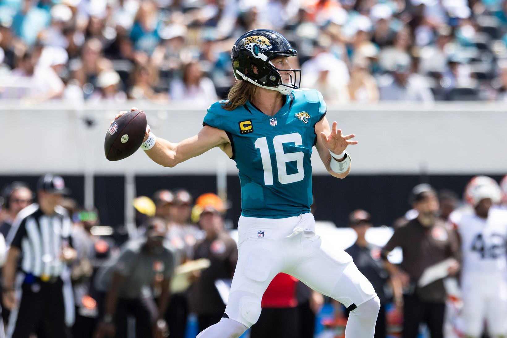 Jacksonville Jaguars quarterback Trevor Lawrence (16), wearing a captain's patch, winds up to throw a pass with an official and a Cleveland Browns player in the background.