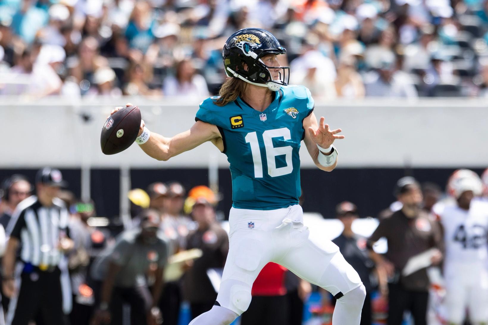Jacksonville Jaguars quarterback Trevor Lawrence (16), wearing a captain's patch, winds up to throw a pass with an official and a Cleveland Browns player in the background.