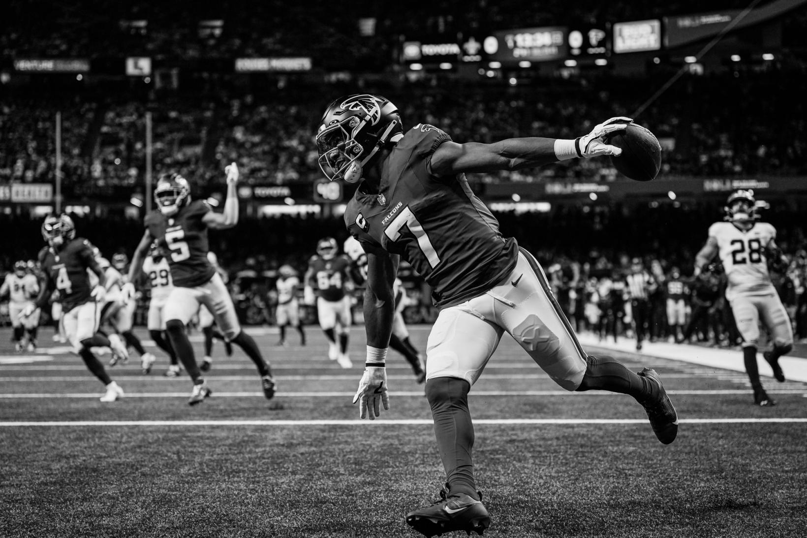 Black-and-white photo of Atlanta Falcons running back Bijan Robinson (7) holding the ball out with one arm as he celebrates a touchdown at Caesars Superdome, with teammates running toward him