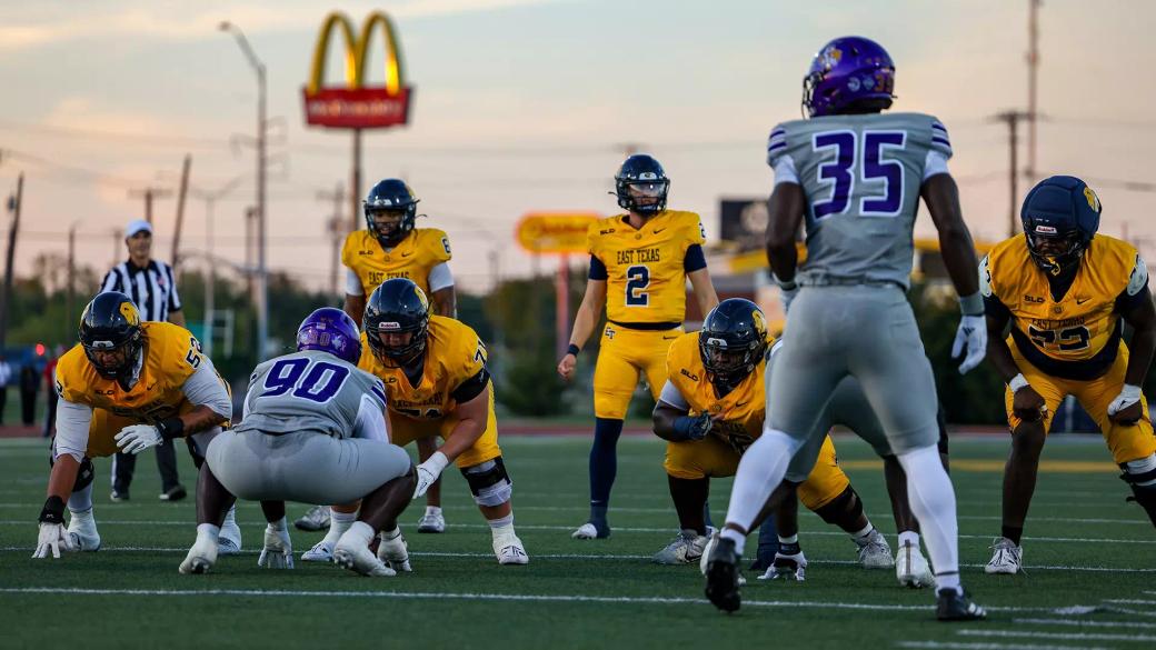 East Texas A&M quarterback Reese Mooney, wearing No. 2 in gold, lines up in the shotgun as Prairie View A&M defenders in gray wait at the line at Memorial Stadium in Commerce