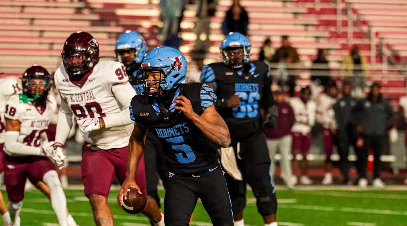 A Delaware State ball carrier in a black jersey and light blue helmet runs with the football as North Carolina Central defenders in white and maroon pursue in Dover