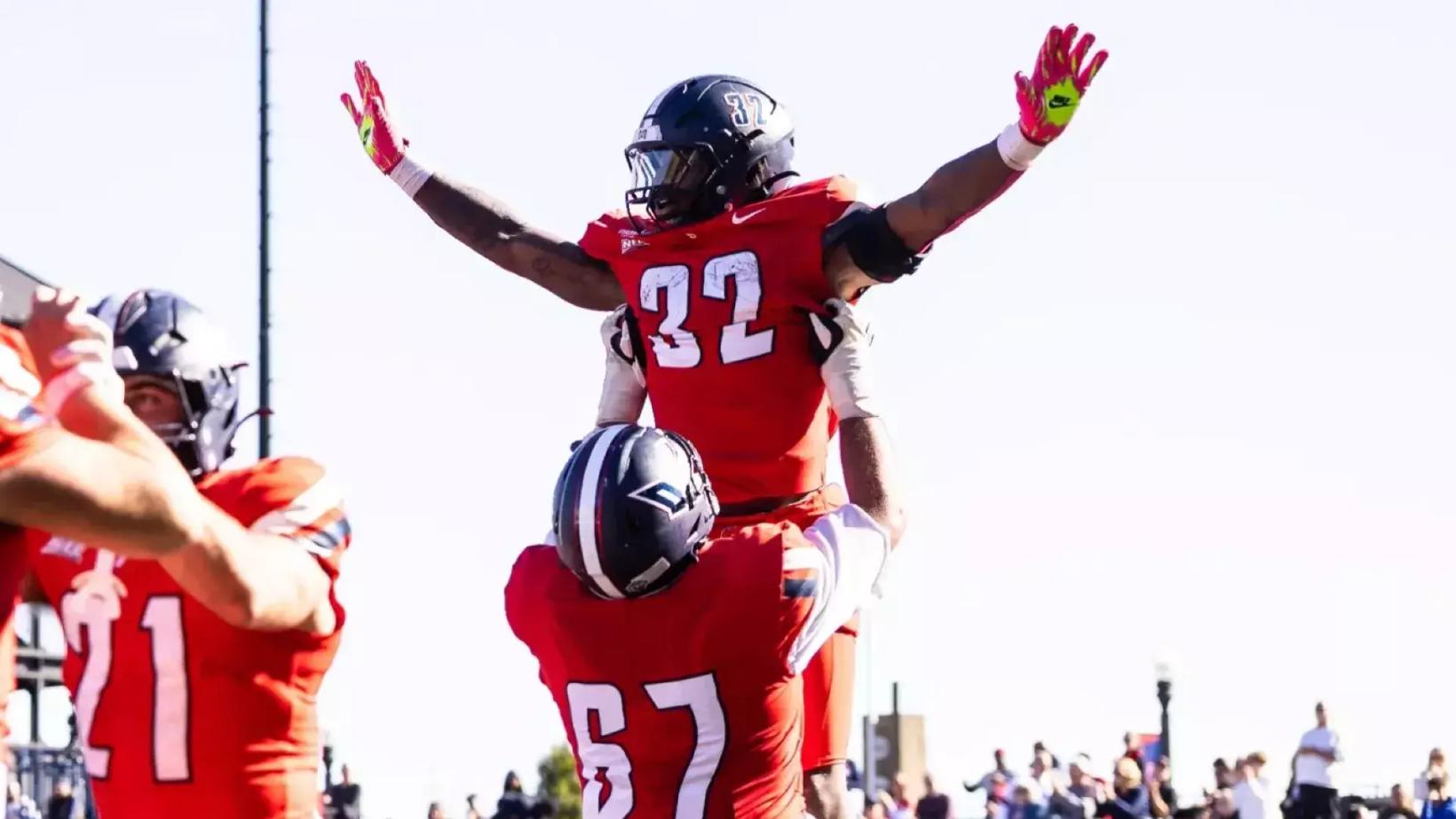 Duquesne running back Ness Davis, No. 32 in a red jersey, raises his arms as teammate No. 67 lifts him to celebrate a touchdown run against Wagner at Rooney Field on Oct. 24, 2026