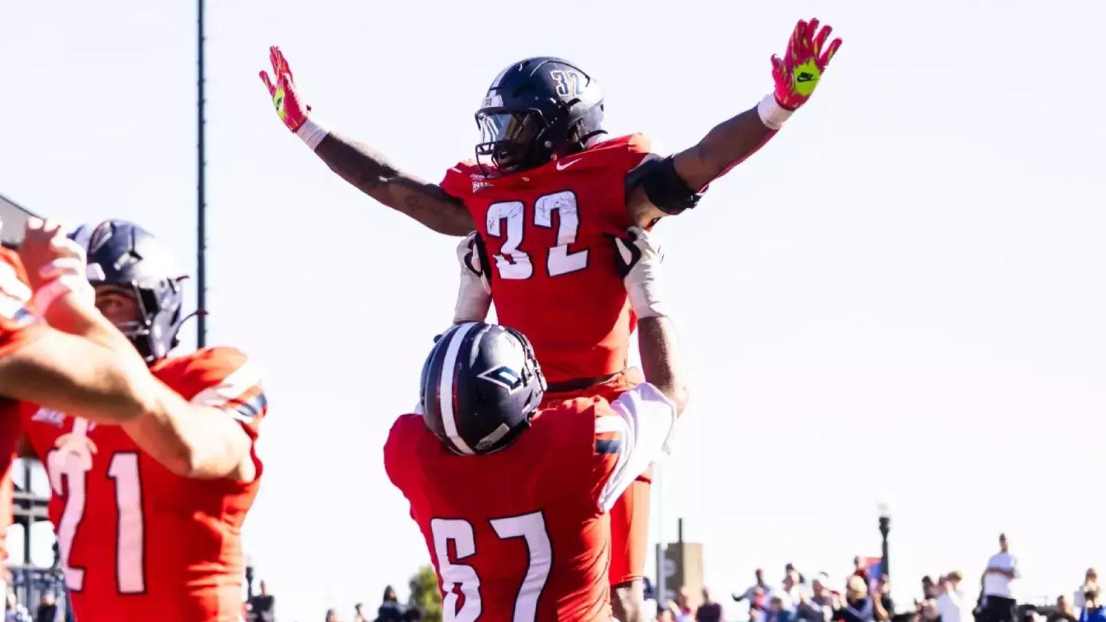 Duquesne running back Ness Davis, No. 32 in a red jersey, raises his arms as teammate No. 67 lifts him to celebrate a touchdown run against Wagner at Rooney Field on Oct. 24, 2026