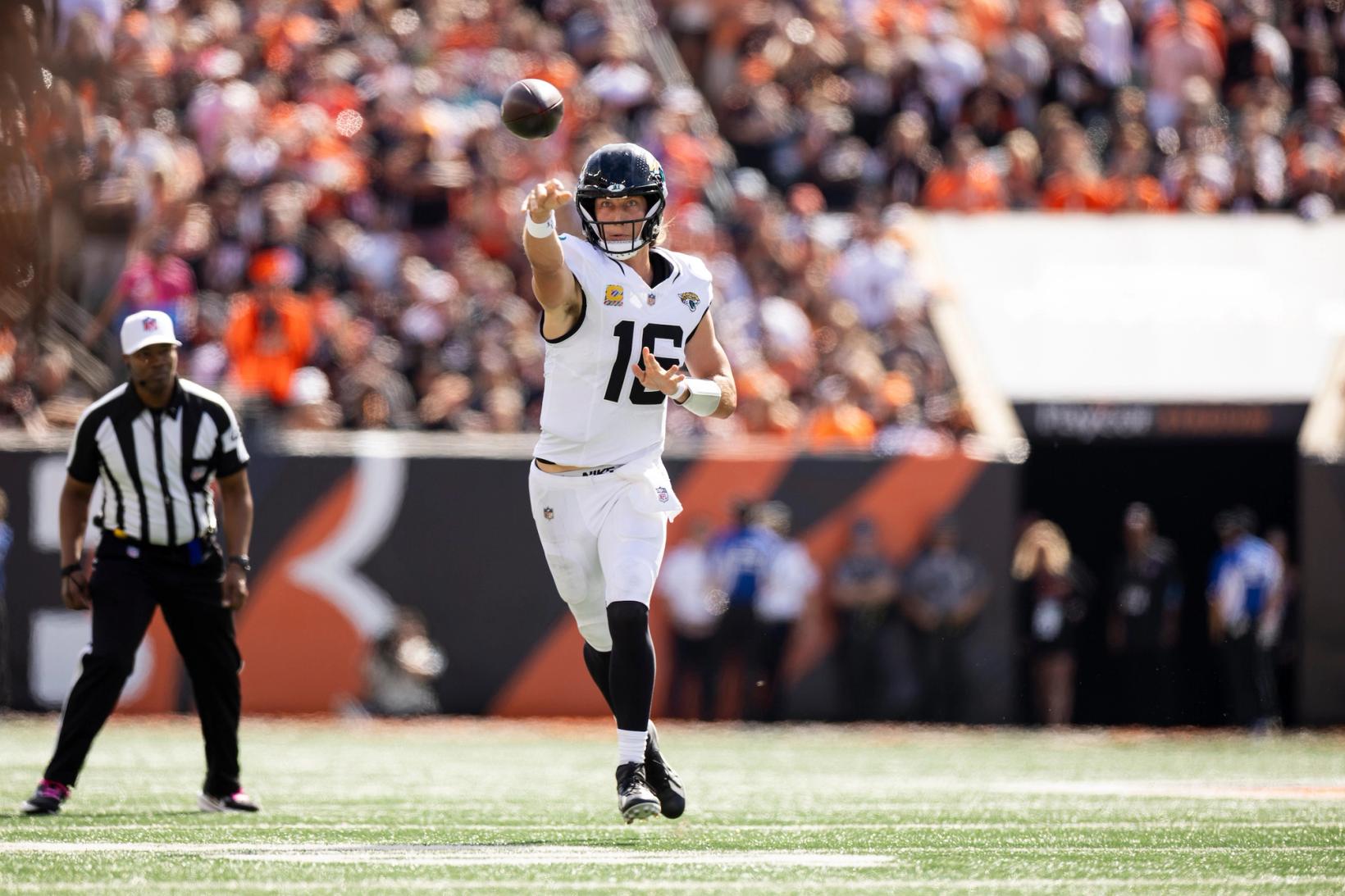 Jacksonville Jaguars quarterback Trevor Lawrence (16) releases a pass near the sideline as an official looks on, with Bengals stripes on the wall behind him.