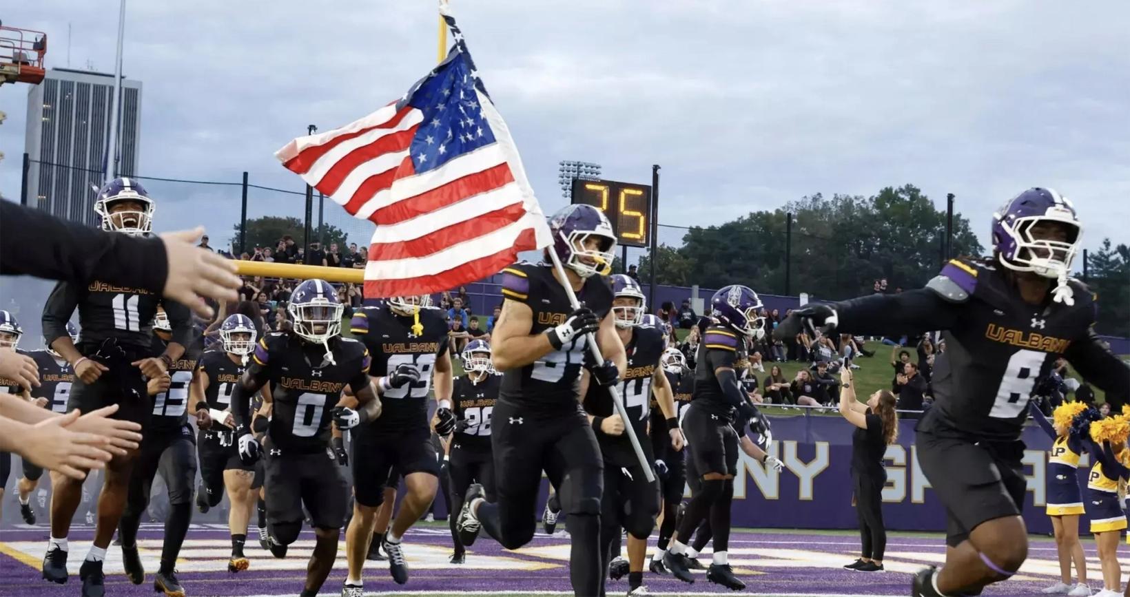 A UAlbany football player carries the American flag onto the field at Casey Stadium, surrounded by teammates, before the 2026 opener; used with UAlbany's 45-35 win over Campbell on Oct. 31, 2026