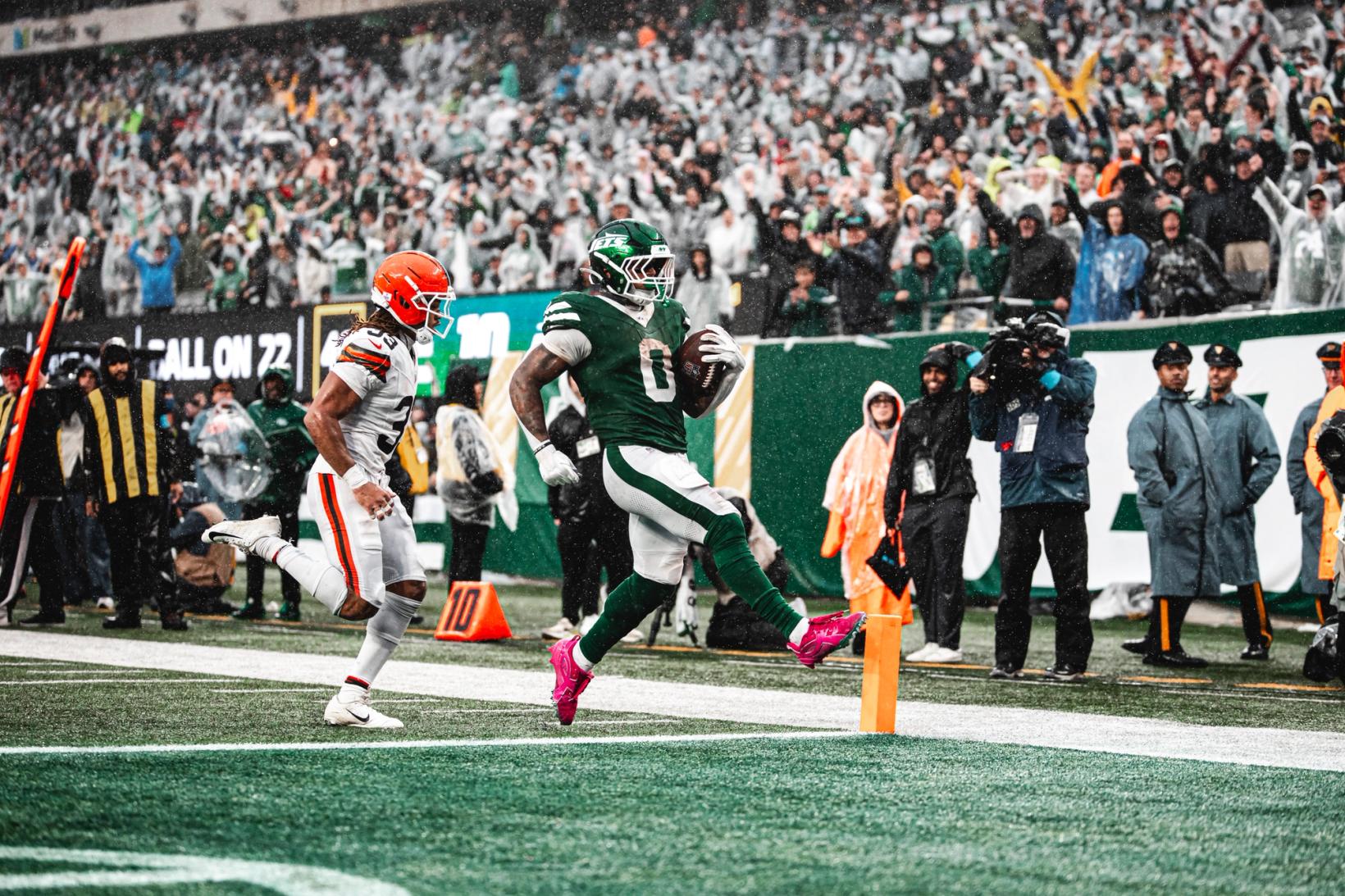 New York Jets ball carrier Braelon Allen (0) crosses the goal line beside the pylon ahead of a chasing Cleveland Browns defender as fans in rain ponchos cheer at MetLife Stadium.