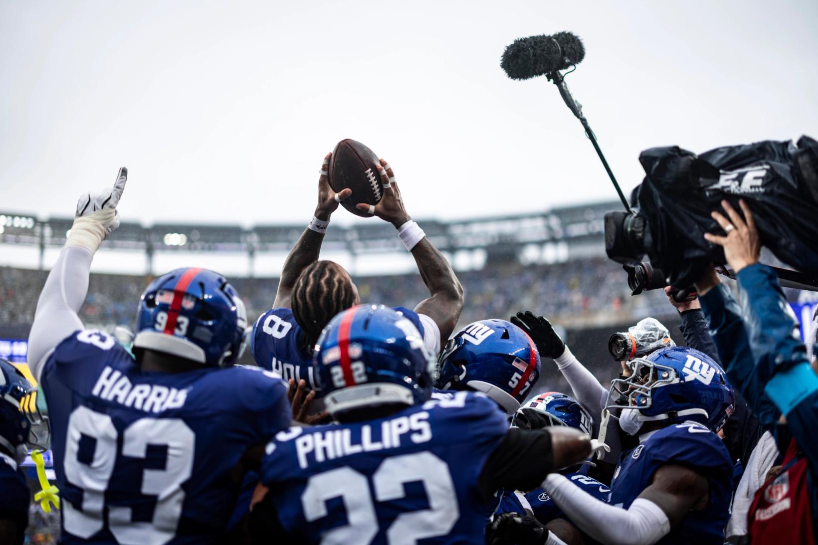 New York Giants safety Jevon Holland (8) holds the football overhead as teammates, including cornerback Dru Phillips (22), crowd around him under gray skies at MetLife Stadium