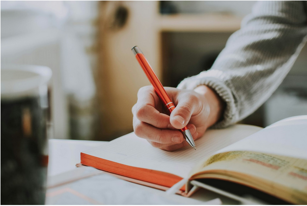 Person in gray sweater writing in an open notebook with a red pen