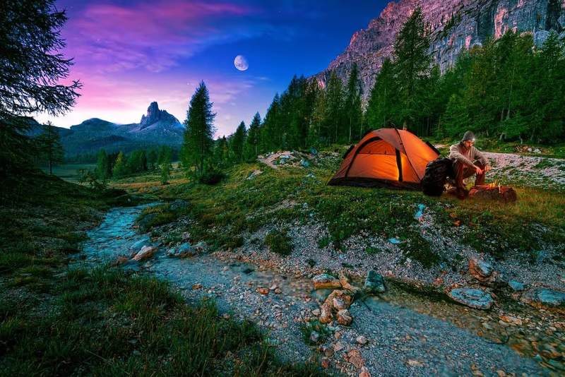 Homme en bivouac au bord d’un ruisseau en montagne, sous une tente éclairée au crépuscule