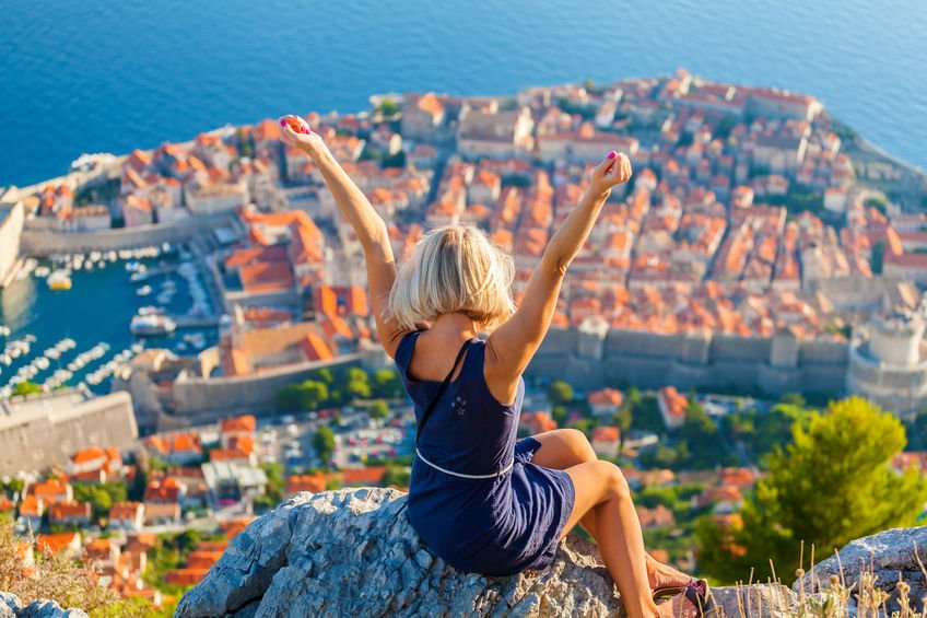 Une femme assise sur un rocher, les bras levés en signe de joie, avec une vue panoramique sur la ville fortifiée de Dubrovnik en Croatie en arrière-plan, illustrant l'émerveillement et la beauté des paysages croates.
