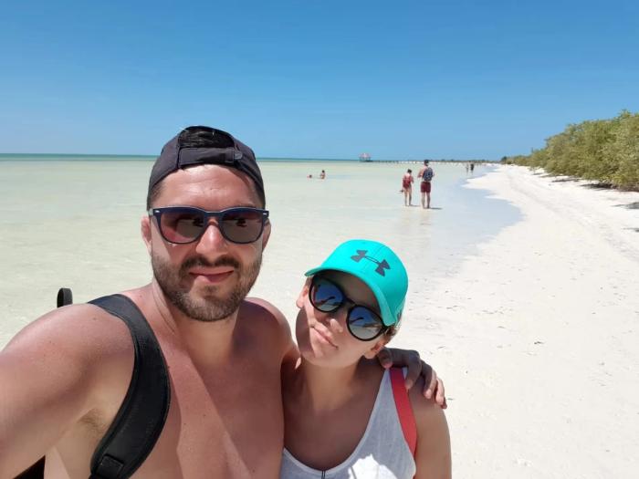 Selfie de Cyril et Maud sur la plage de Holbox, avec une mer turquoise en arrière-plan et des visiteurs profitant du sable blanc.