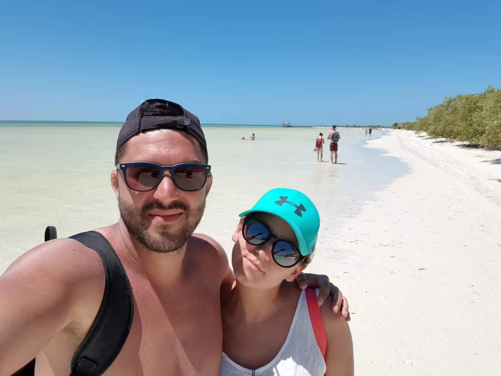 Selfie de Cyril et Maud sur la plage de Holbox, avec une mer turquoise en arrière-plan et des visiteurs profitant du sable blanc.