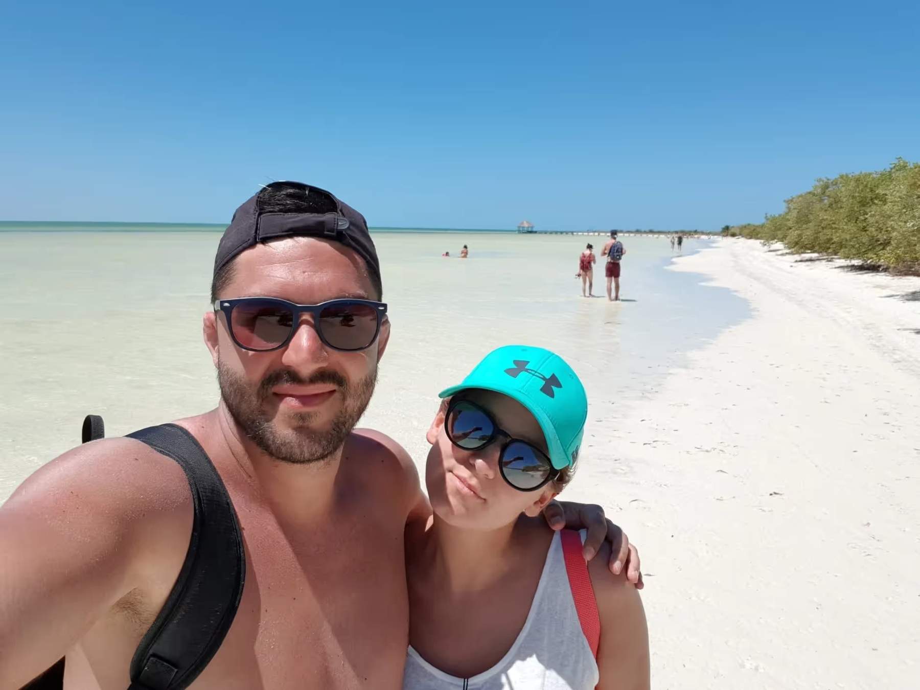 Selfie de Cyril et Maud sur la plage de Holbox, avec une mer turquoise en arrière-plan et des visiteurs profitant du sable blanc.