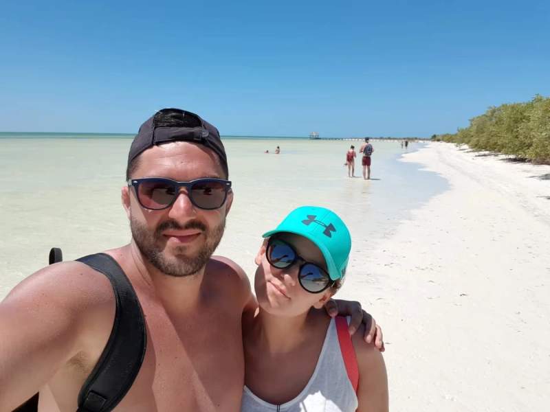 Selfie de Cyril et Maud sur la plage de Holbox, avec une mer turquoise en arrière-plan et des visiteurs profitant du sable blanc.