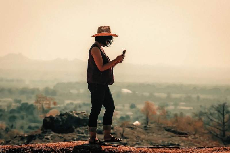 Une femme portant un chapeau, utilisant son téléphone portable en plein air avec un paysage flou en arrière-plan.