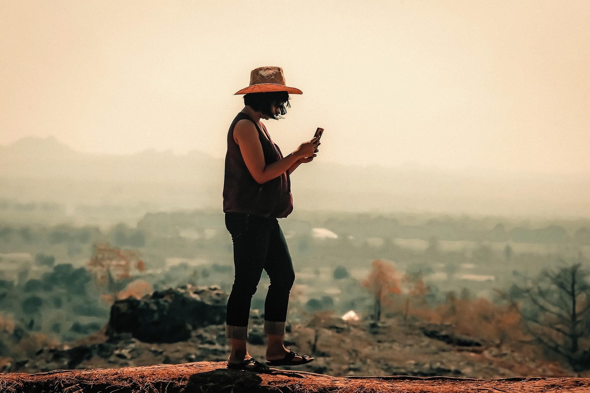 Une femme portant un chapeau, utilisant son téléphone portable en plein air avec un paysage flou en arrière-plan.