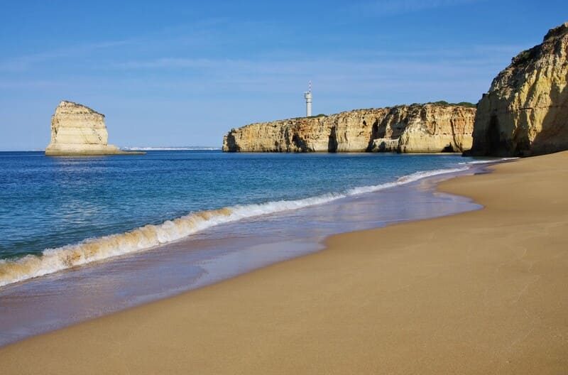 Une plage de sable doré bordée de falaises calcaires, avec des vagues douces et une mer bleu azur sous un ciel dégagé.