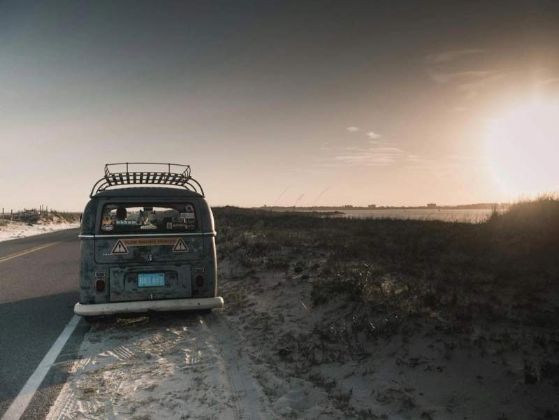 Un van vintage garé sur le bord d'une route côtière, avec le soleil se couchant sur une plage déserte en arrière-plan.