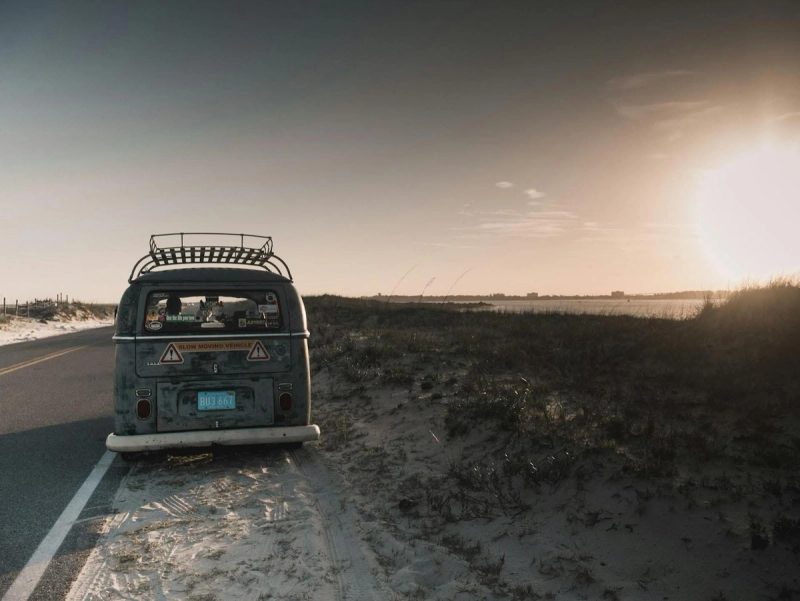 Un van vintage garé sur le bord d'une route côtière, avec le soleil se couchant sur une plage déserte en arrière-plan.