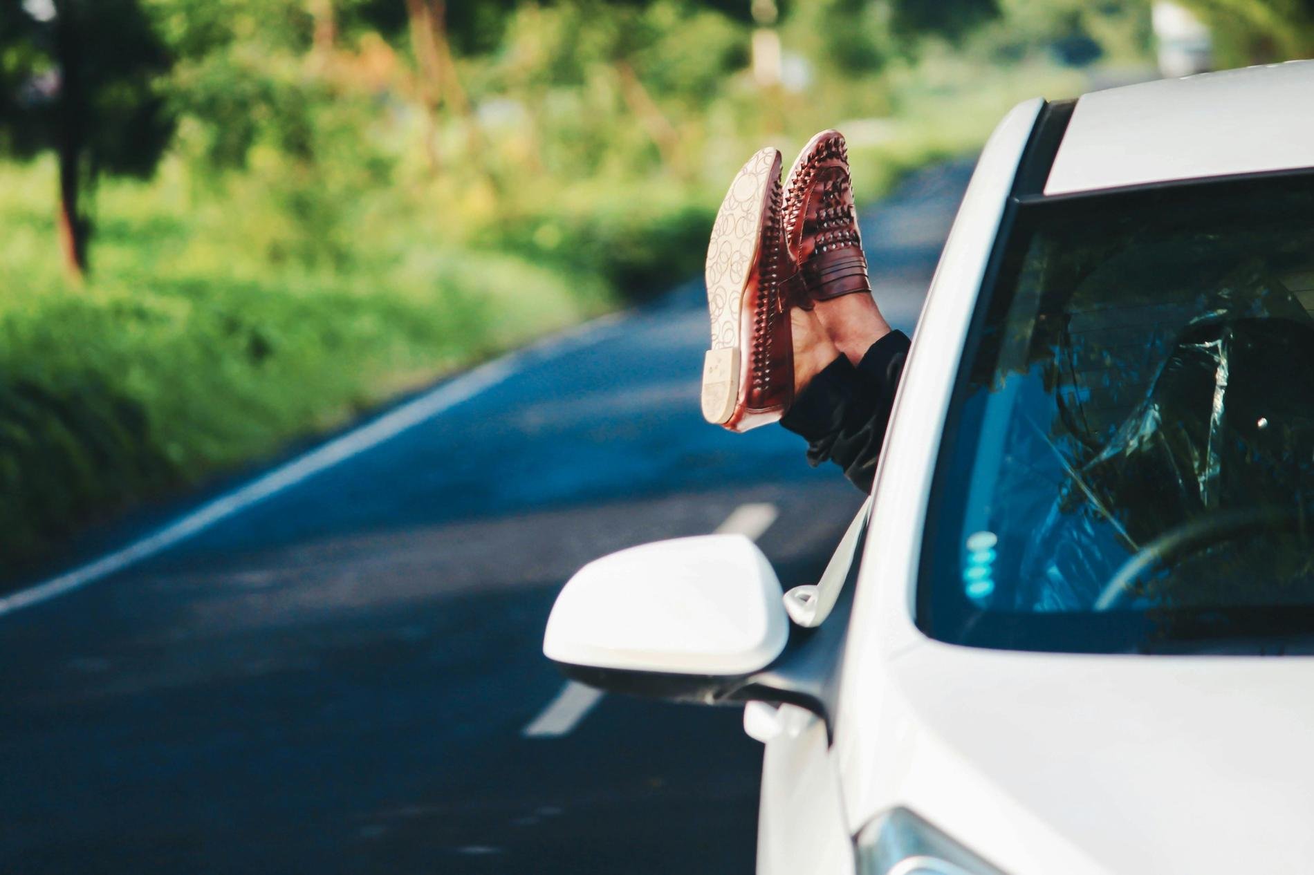 Des pieds chaussés de mocassins sortent par la fenêtre d'une voiture blanche, roulant sur une route entourée de verdure, suggérant une ambiance de détente et de road trip. ​