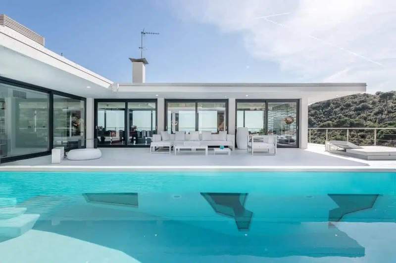 Villa contemporaine avec grande terrasse blanche et piscine turquoise, située à Sitges, offrant une vue dégagée sur la nature environnante.