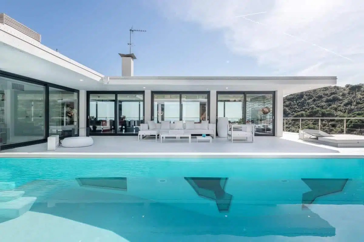 Villa contemporaine avec grande terrasse blanche et piscine turquoise, située à Sitges, offrant une vue dégagée sur la nature environnante.