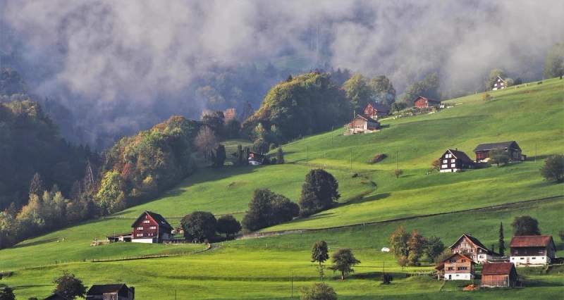 Vue panoramique sur une vallée suisse verdoyante, parsemée de chalets traditionnels et entourée de collines embrumées, capturant l'essence d'un paysage alpin paisible.