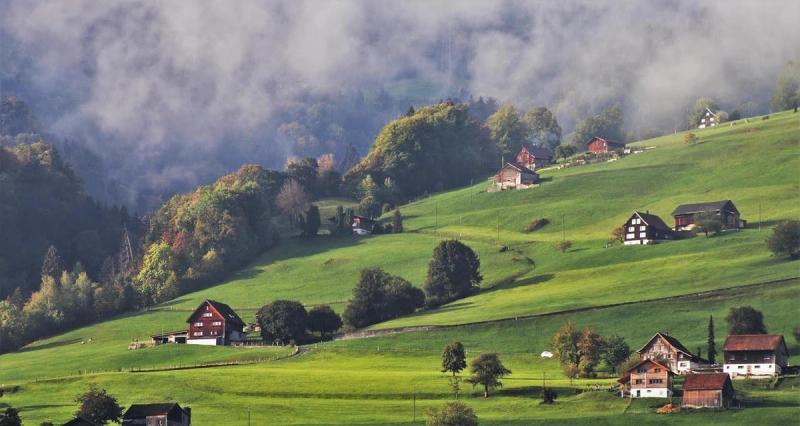 Vue panoramique sur une vallée suisse verdoyante, parsemée de chalets traditionnels et entourée de collines embrumées, capturant l'essence d'un paysage alpin paisible.