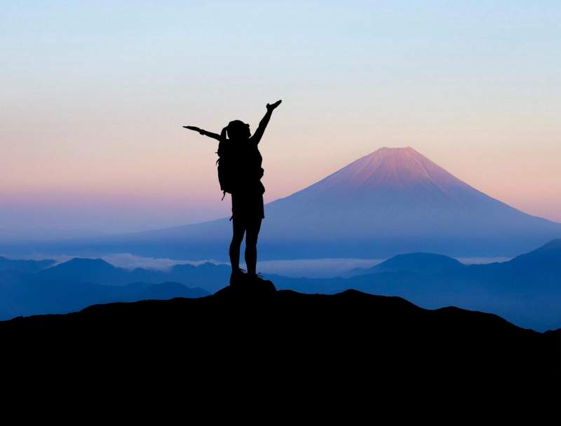 Silhouette d’un voyageur célébrant au sommet d’une montagne avec le Mont Fuji en arrière-plan, au lever du soleil.