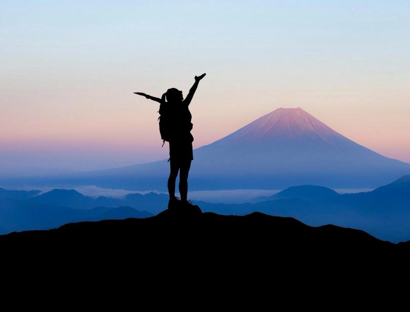 Silhouette d’un voyageur célébrant au sommet d’une montagne avec le Mont Fuji en arrière-plan, au lever du soleil.