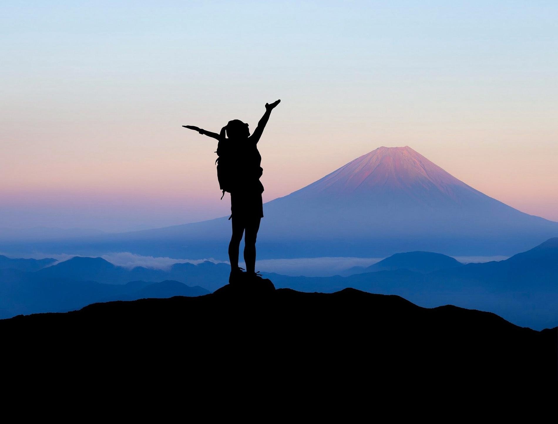 Silhouette d’un voyageur célébrant au sommet d’une montagne avec le Mont Fuji en arrière-plan, au lever du soleil.
