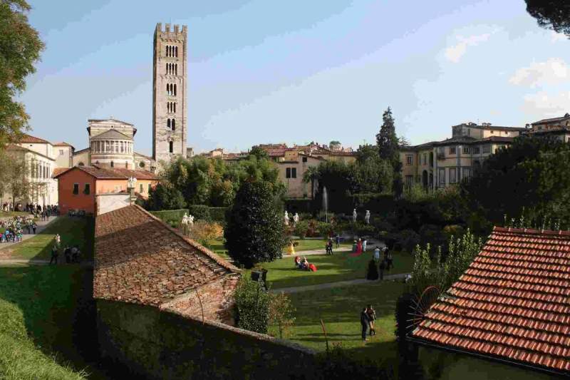 Vue générale de Lucca avec des jardins, des bâtiments historiques et une tour en arrière-plan sous un ciel bleu.