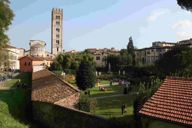 Vue générale de Lucca avec des jardins, des bâtiments historiques et une tour en arrière-plan sous un ciel bleu.