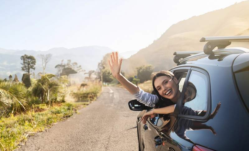Jeune femme souriante passant la tête par la fenêtre d'une voiture sur une route de montagne lors d'un road trip en Europe