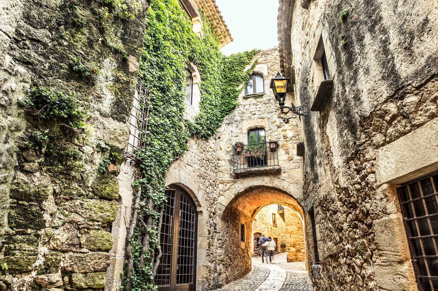 Ruelle pavée étroite dans le village médiéval de Pals, avec des murs en pierre couverts de lierre et des fenêtres en fer forgé.