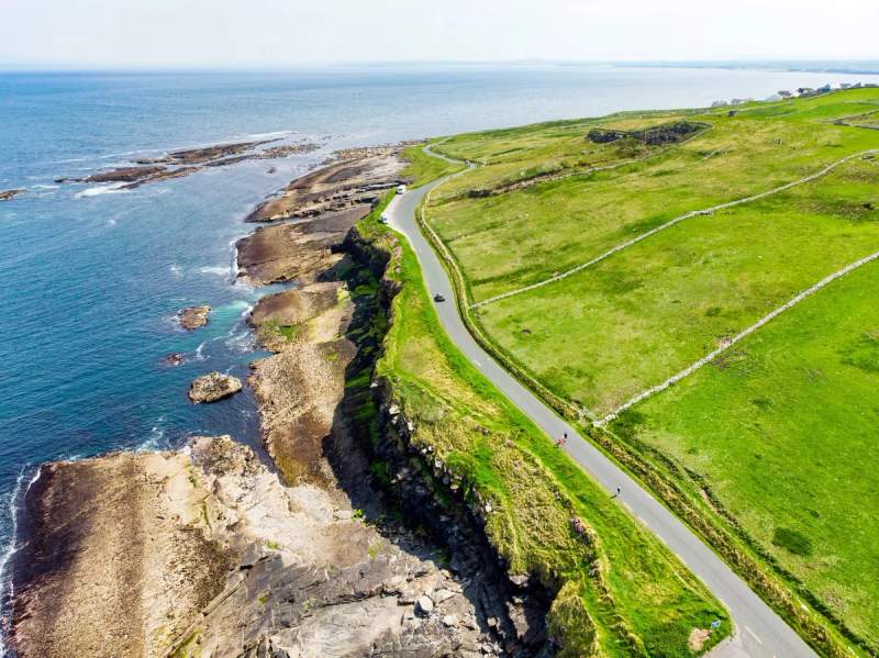 Vue aérienne d’une route côtière en Irlande, longeant des falaises verdoyantes et bordant l’océan.